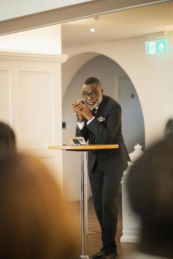 Tunji Idowu in a dark suit, leaning on a standing table with hands clasped, listening