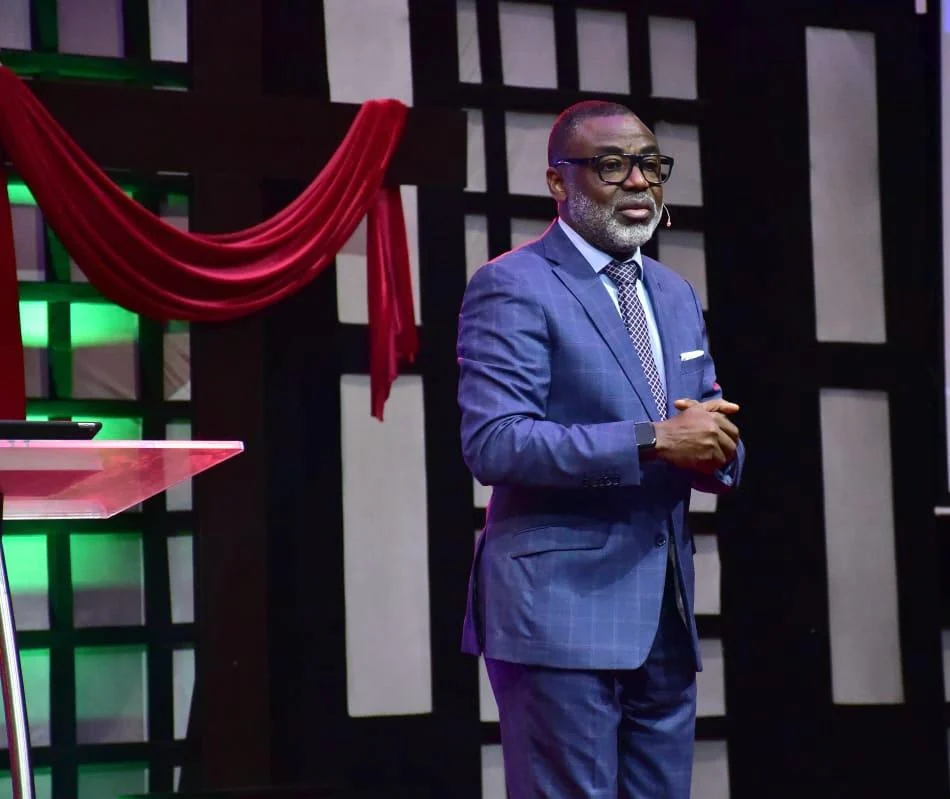 Tunji Idowu speaking on a stage beside a red drape, hands clasped