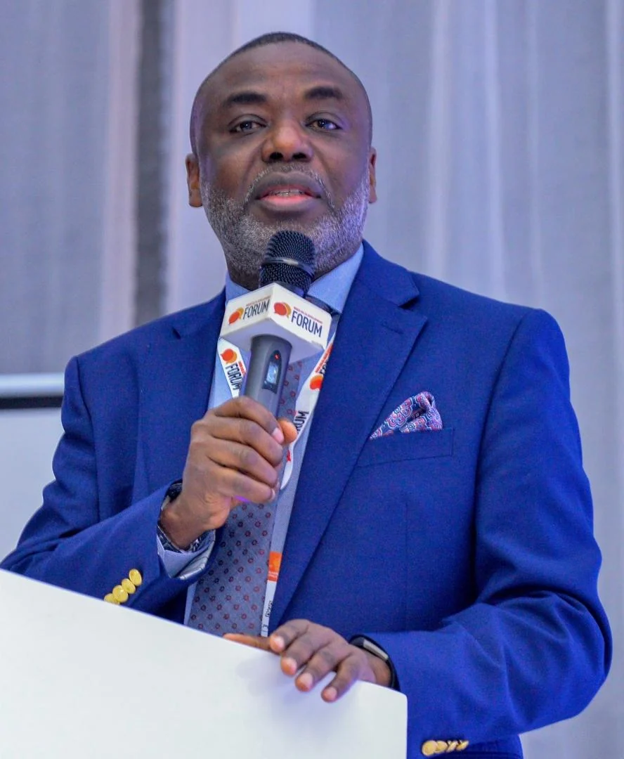 Tunji Idowu in a blue suit speaking into a Forum-branded microphone at a lectern