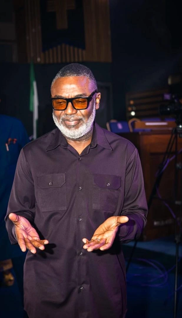 Tunji Idowu smiling with his palms open, wearing amber-tinted glasses and a dark shirt, in a dimly lit hall