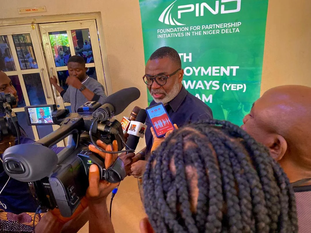 Tunji Idowu answering questions from reporters holding microphones and a video camera, in front of a Foundation for Partnership Initiatives in the Niger Delta banner