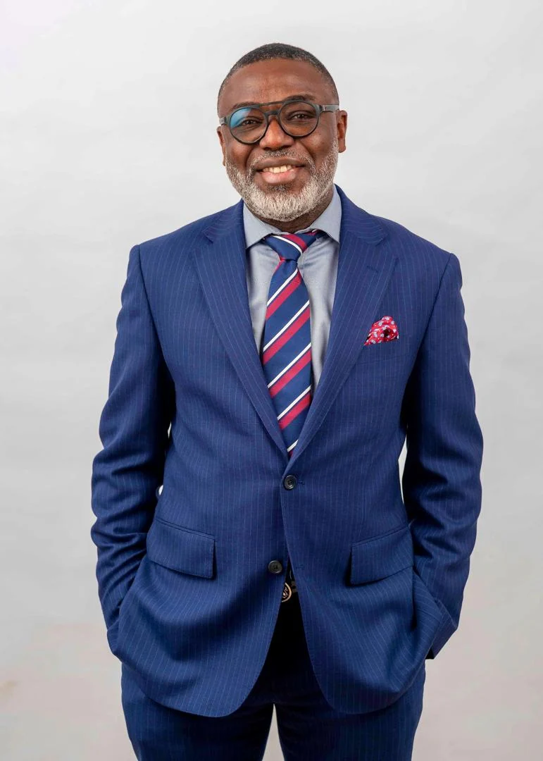 Tunji Idowu, smiling, in a blue pinstripe suit against a plain light background