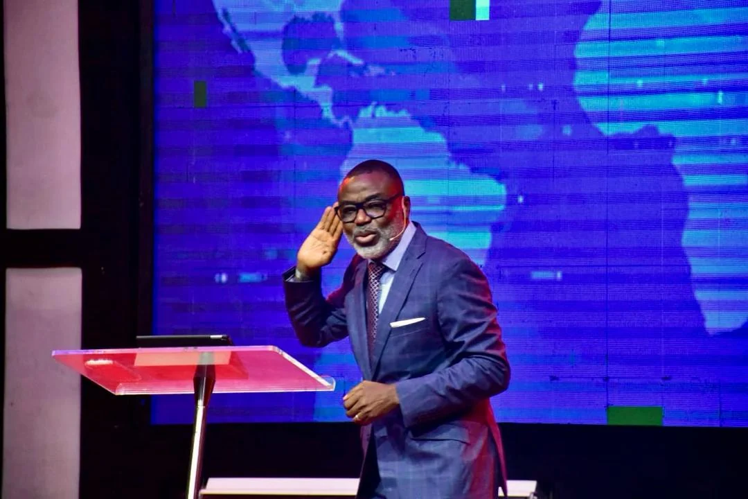 Tunji Idowu on stage with a hand raised to one ear, in front of a projected world map