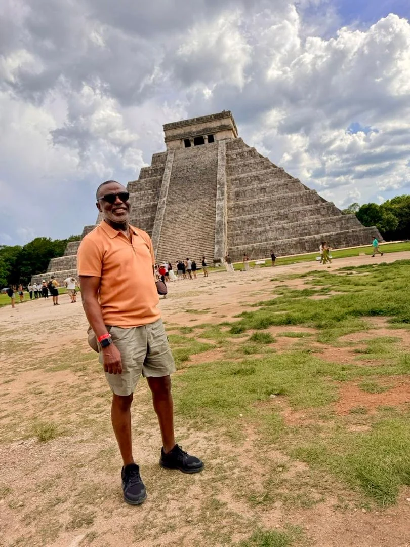 Tunji Idowu smiling in front of a large stepped stone pyramid under a cloudy sky