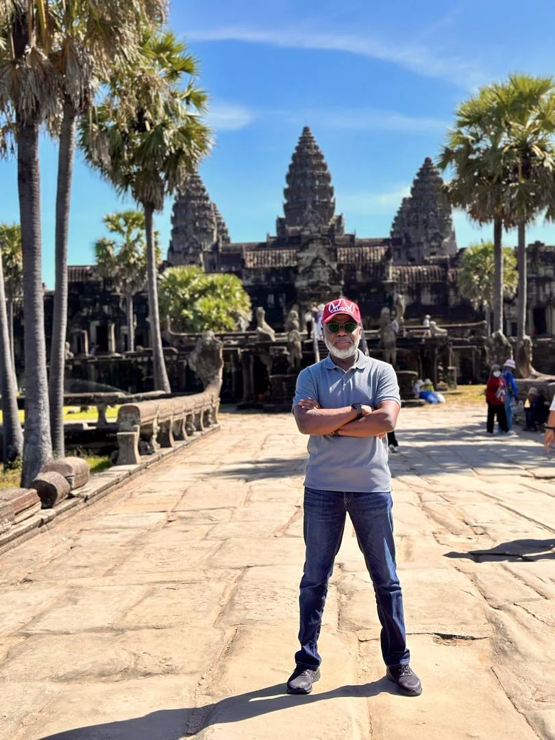 Tunji Idowu standing with arms folded on a stone walkway in front of an ancient temple with carved towers and palm trees